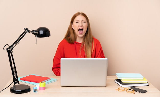 Mulher jovem estudante em um local de trabalho com um laptop gritando para a frente com a boca aberta