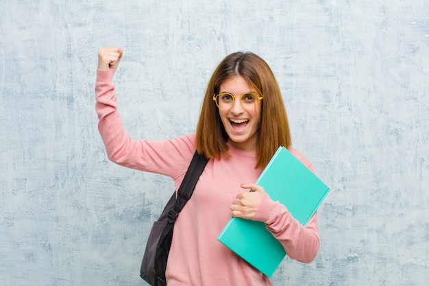 Mulher jovem estudante gritando triunfante, parecendo vencedor animado, feliz e surpreso, comemorando contra o fundo da parede grunge