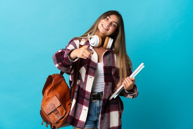 Mulher jovem estudante isolada em azul apontando para a frente com uma expressão feliz