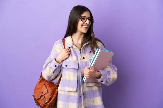 Mulher jovem estudante isolada em um fundo roxo, olhando para o lado e sorrindo