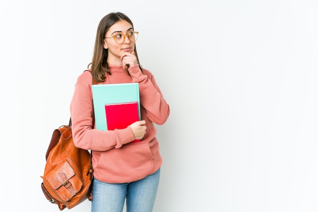 Mulher jovem estudante isolada na parede branca olhando de soslaio com expressão duvidosa e cética