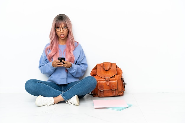 Mulher jovem estudante mestiça de cabelo rosa sentada no chão, isolada no fundo branco, surpresa e enviando uma mensagem