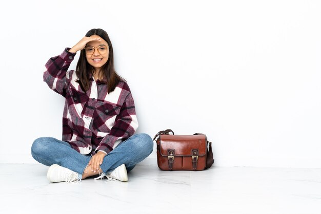 Mulher jovem estudante sentada no chão saudando com a mão com expressão feliz