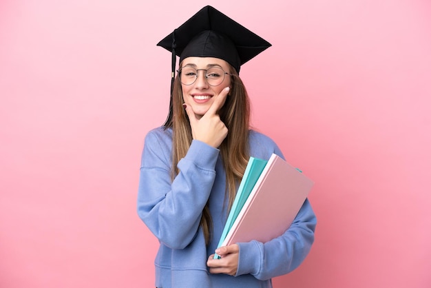 Mulher jovem estudante usando um chapéu de pós-graduação isolado em fundo rosa feliz e sorridente
