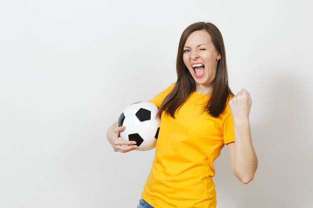 Mulher jovem europeia bonita, fã de futebol ou jogador de uniforme amarelo, segurando uma bola de futebol, mostrando o gesto do vencedor isolado no fundo branco. Esporte jogo saúde futebol, conceito de estilo de vida saudável.