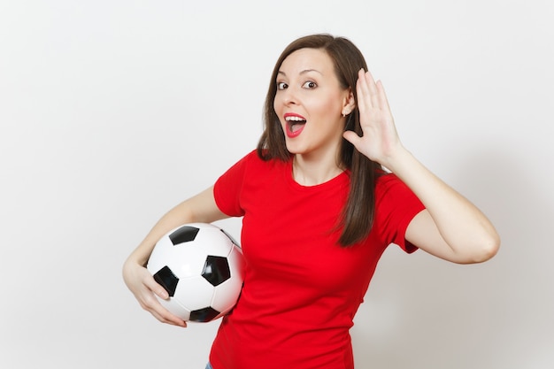 Mulher jovem europeia bonita, fã de futebol ou jogador de uniforme vermelho bisbilhotar, ouvir o gesto, segurar uma bola de futebol isolada no fundo branco. Esporte joga futebol, saúde, conceito de estilo de vida saudável.