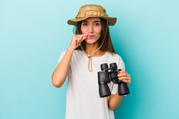 Mulher jovem explorador caucasiana segurando binóculos isolados em um fundo azul com os dedos nos lábios, mantendo um segredo.