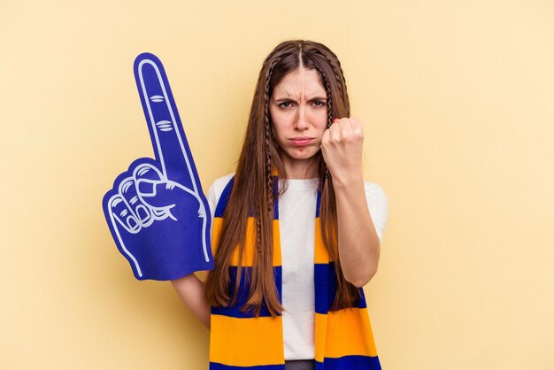 Mulher jovem fã de esportes isolada em fundo amarelo, mostrando o punho para a câmera, expressão facial agressiva.