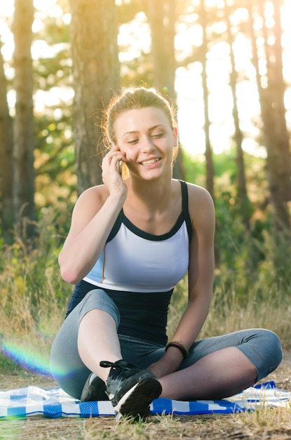 Foto mulher jovem falando no telefone móvel enquanto se exercita contra árvores na floresta