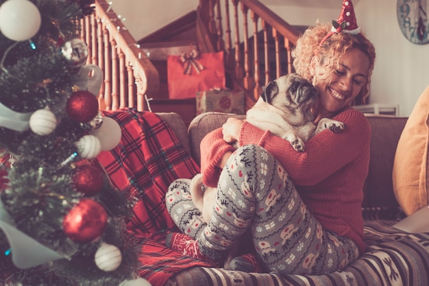 Mulher jovem feliz abraçando o cachorro de estimação e celebrando o Natal. Mulher caucasiana e cachorro pug se divertindo no Natal em casa. Mulher alegre abraçando cachorro no sofá e comemorando o natal