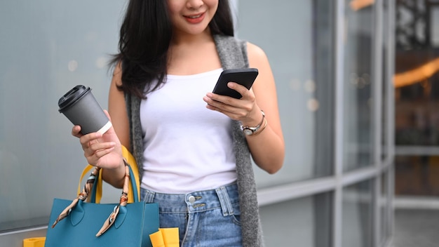 Mulher jovem feliz andando por uma rua no distrito comercial com suas sacolas de compras e usando telefone inteligente