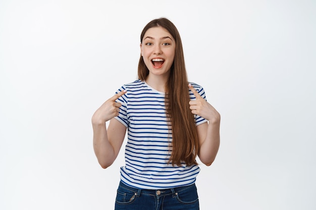 Mulher jovem feliz apontando para si mesma e rindo, sorrindo satisfeita, em pé no branco