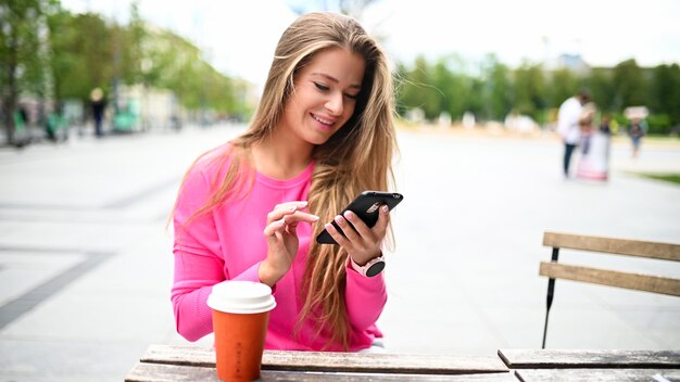 Mulher jovem feliz bebendo um café ao ar livre enquanto usa seu smartphone