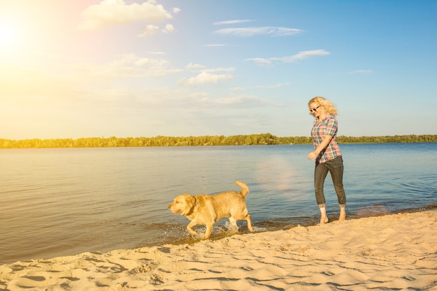 Mulher jovem feliz caminhando em uma praia com seu golden retriever no pôr do sol