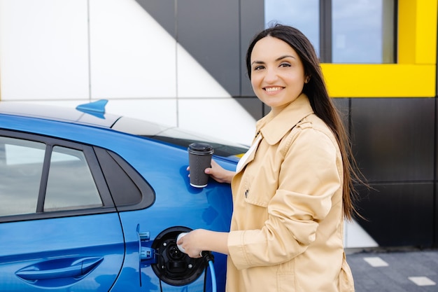 Mulher jovem feliz carregando bateria de automóvel de pequena estação pública perto de carro elétrico bebendo café