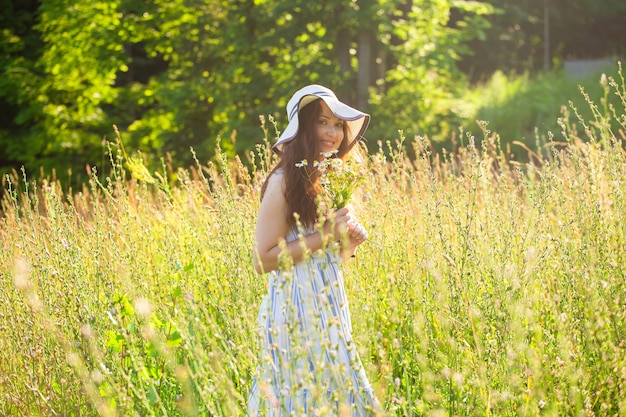 Mulher jovem feliz com cabelo comprido usando chapéu e vestido puxando as mãos em direção às plantas enquanto caminha