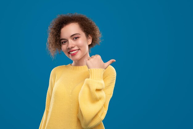 Mulher jovem feliz com cabelo encaracolado, vestindo um suéter amarelo apontando para longe com o polegar contra um fundo azul com um espaço vazio