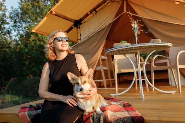 Mulher jovem feliz com cachorro welsh corgi pembroke relaxando em glamping no dia de verão barraca de acampamento de luxo para recreação ao ar livre e recreação conceito de estilo de vida