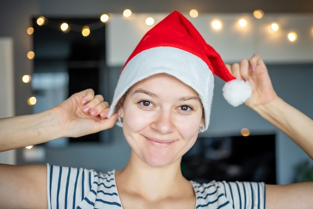 Mulher jovem feliz com chapéu de Papai Noel sorrindo para as luzes decoradas em casa