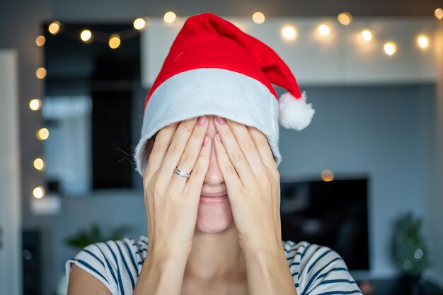 Mulher jovem feliz com chapéu de Papai Noel sorrindo para as luzes decoradas em casa