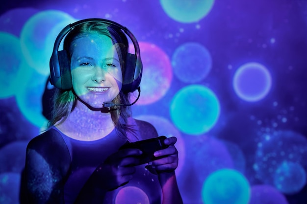 Mulher jovem feliz com fone de ouvido olhando para a câmera com um sorriso e pressionando botões no gamepad enquanto joga videogame sob esportes coloridos de projeção de luz
