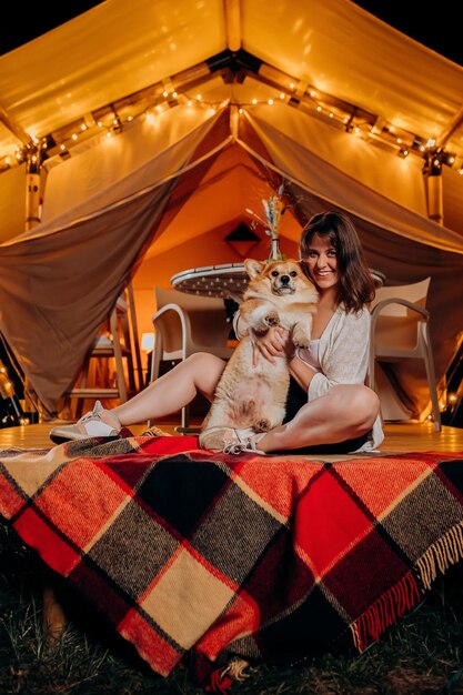 Mulher jovem feliz com seu cachorro Welsh Corgi Pembroke relaxando em glamping na noite de verão perto da fogueira aconchegante Barraca de acampamento de luxo para recreação ao ar livre e recreação Conceito de estilo de vida