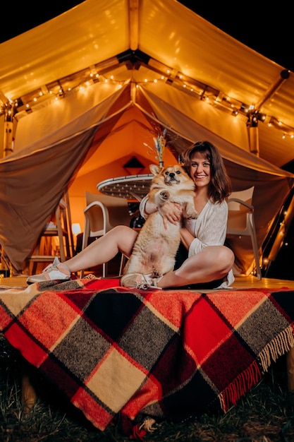 Mulher jovem feliz com seu cachorro welsh corgi pembroke relaxando em glamping na noite de verão perto da fogueira aconchegante barraca de acampamento de luxo para recreação ao ar livre e recreação conceito de estilo de vida