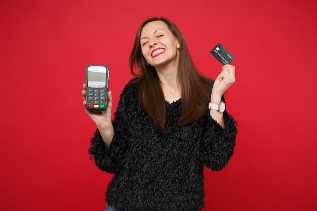 Mulher jovem feliz com suéter de pele preta segurando o terminal de pagamento do banco moderno sem fio para processar e adquirir pagamentos com cartão de crédito isolados sobre fundo vermelho. emoções sinceras de pessoas, conceito de estilo de vida.