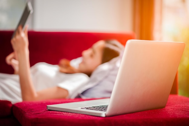Mulher jovem feliz com tablet deitado no sofá e tem um laptop.