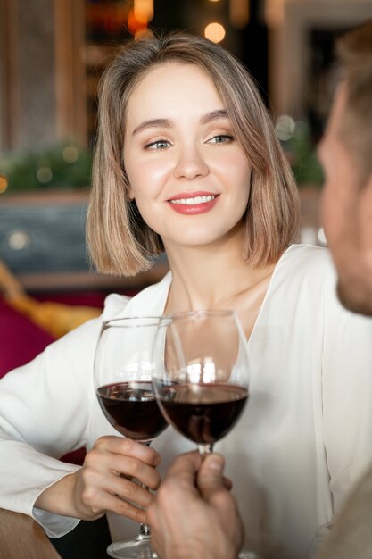 Mulher jovem feliz com um copo de vinho tinto olhando para o namorado com um sorriso cheio de dentes durante o brinde por seu relacionamento no restaurante