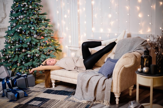 Mulher jovem feliz com um presente deitado perto da árvore de Natal Cor branca prateada no fundo do sofá Feliz Natal e Ano Novo Momentos atmosféricos