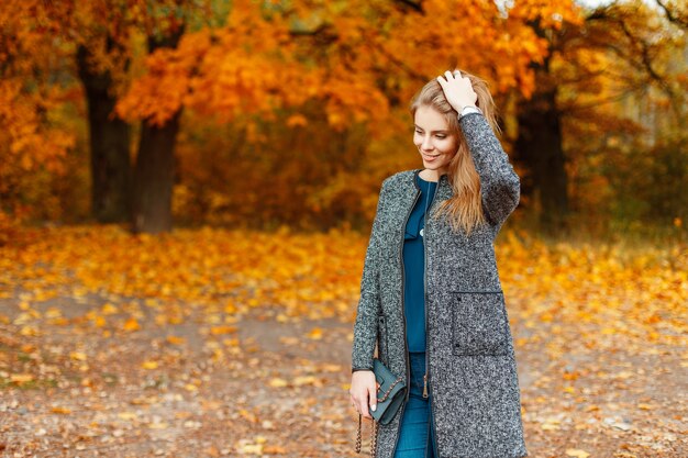 Mulher jovem feliz com um sorriso em roupas da moda de outono se passando perto de árvores com folhagem amarela