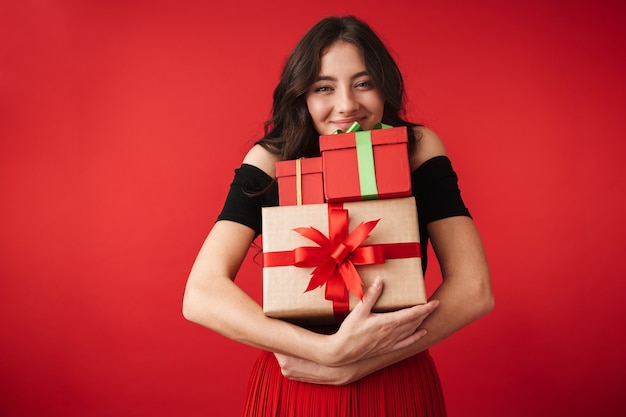 Mulher jovem feliz com um vestido isolado sobre o vermelho, segurando uma pilha de caixas de presente