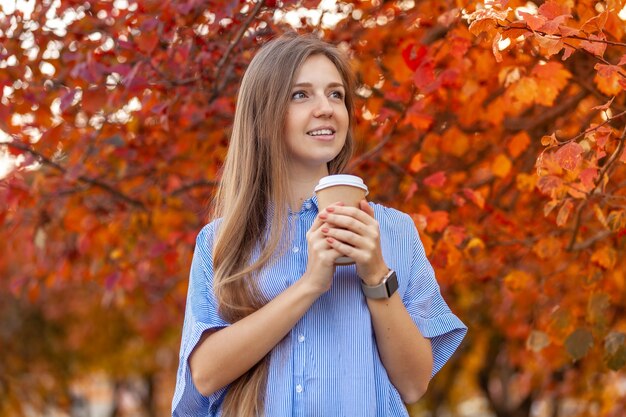 Mulher jovem feliz com uma xícara de café para ir no outono vermelho árvores