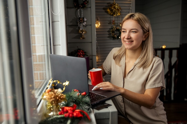 Mulher jovem feliz com uma xícara de café usando o zoom para parabenizar o feliz natal seus parentes. fazendo videochamadas facetime com laptop em casa.