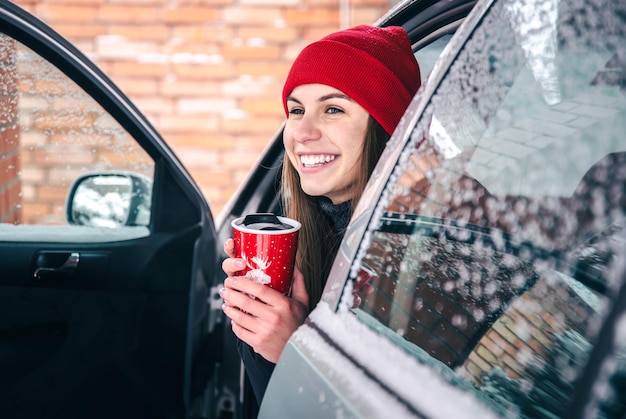 Mulher jovem feliz com uma xícara térmica vermelha sentada em um carro no inverno