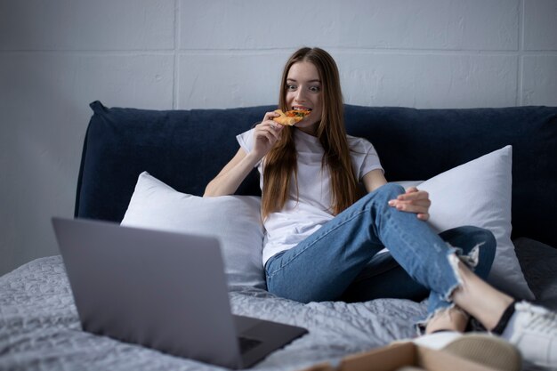 Mulher jovem feliz comendo uma fatia de pizza quente em casa e assistindo filme no laptop