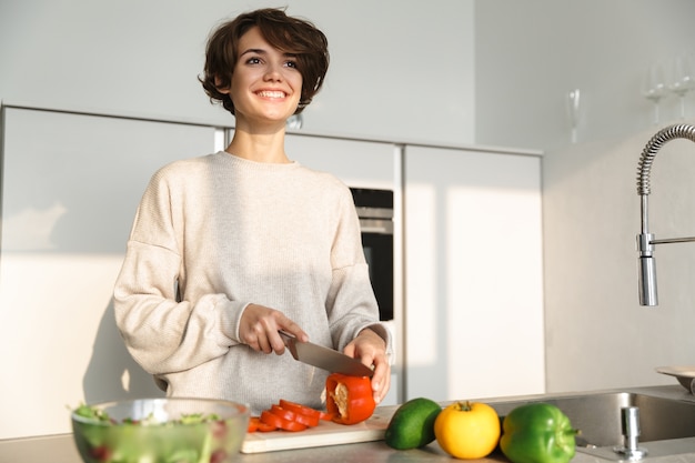 Mulher jovem feliz cozinhando salada fresca na cozinha de casa