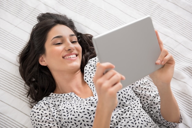 Mulher jovem feliz deitada na cama usando tablet digital