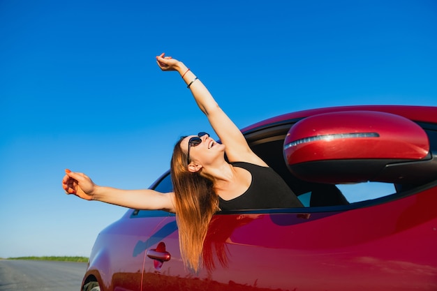 Mulher jovem feliz desfrutar de viagem, metade do corpo pela janela automática aberta e levantar as mãos, usando óculos escuros.