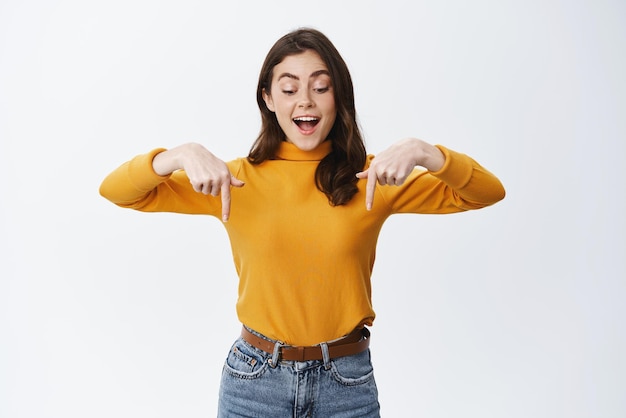 Foto mulher jovem feliz e animada, parecendo hipnotizada e fascinada, apontando os dedos no fundo do espaço vazio para o anúncio mostrando o fundo branco do logotipo