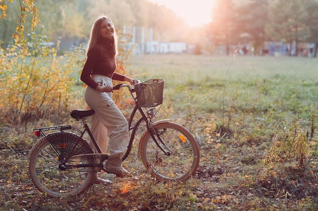 Mulher jovem feliz e ativa andando de bicicleta vintage no parque de outono ao pôr do sol