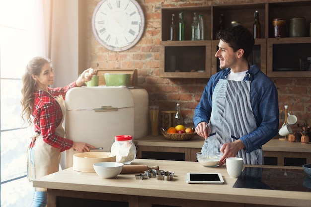 Mulher jovem feliz e homem fazendo torta na cozinha do sótão. Família jovem cozinhando em casa, usando tablet digital.