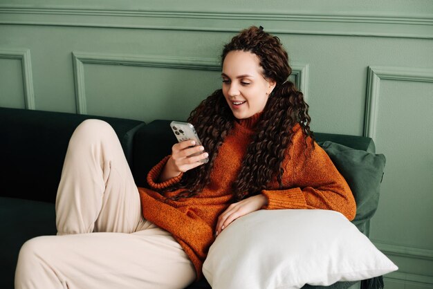 Mulher jovem feliz e relaxada sentada no sofá usando telefone celular senhora sorridente rindo segurando smartphone olhando para o celular gostando de fazer compras de comércio eletrônico on-line em aplicativos móveis ou assistir a vídeos