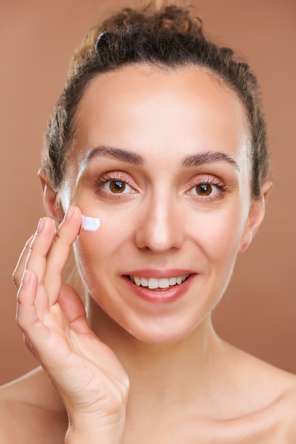 Foto mulher jovem feliz e saudável aplicando creme hidratante no rosto enquanto fica em frente à câmera contra uma parede marrom