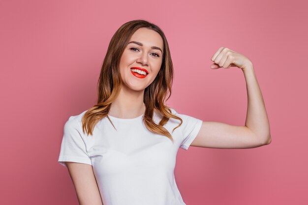 Mulher jovem feliz em camiseta branca sorrindo e mostra seus músculos isolados na parede rosa.
