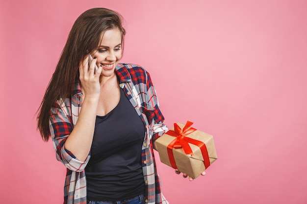 Mulher jovem feliz em casual segurando a caixa de presente e olhando de lado enquanto desfruta sobre fundo rosa, usando o telefone.