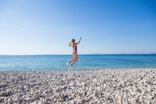 Mulher jovem feliz em estilo casual, pulando na praia de pedra. Relaxante, divertido e aproveite as férias na praia paradisíaca, com céu azul e mar. Garota nas férias de verão.