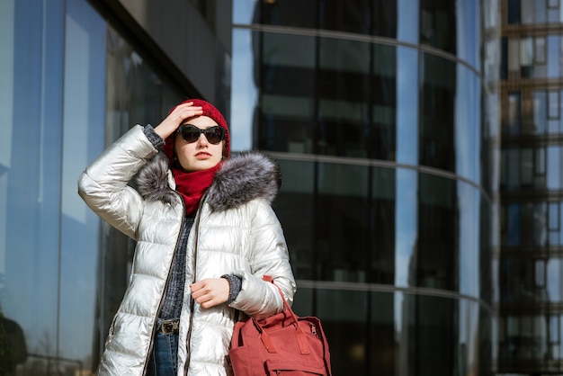 Mulher jovem feliz em óculos de sol com uma jaqueta de inverno com uma mochila perto do edifício. Conceito de viagens