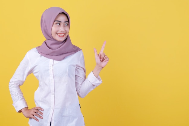 Mulher jovem feliz em pé com o dedo apontando para o lado direito usando um hijab